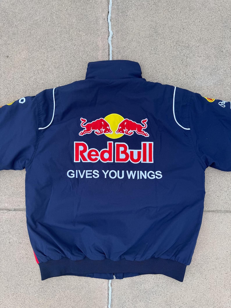 May include: Navy blue bomber jacket featuring the Red Bull logo in red and yellow. The jacket has white accents and the text "GIVES YOU WINGS" in white. The jacket is a sporty style.