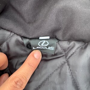 Pu&ograve; includere: Primo piano di un colletto di giacca nero con un motivo trapuntato. Il logo Lexus &egrave; visualizzato su un'etichetta nera. L'etichetta &egrave; rettangolare con testo bianco e un logo bianco.