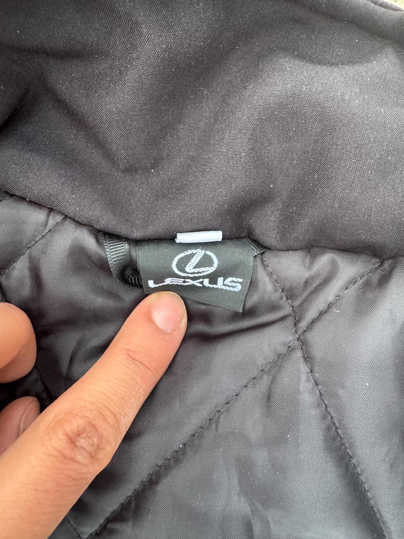 Pu&ograve; includere: Primo piano di un colletto di giacca nero con un motivo trapuntato. Il logo Lexus &egrave; visualizzato su un'etichetta nera. L'etichetta &egrave; rettangolare con testo bianco e un logo bianco.
