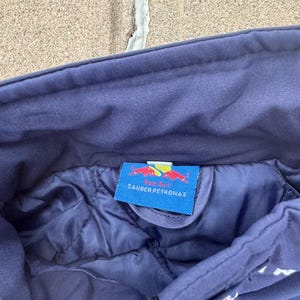 May include: Close-up of a navy blue jacket interior. A tag with the text "SAUBER PETRONAS" and a red bull logo is visible. The jacket has a quilted lining and a zipper.