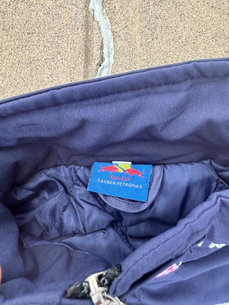 May include: Close-up of a navy blue jacket interior. A tag with the text "SAUBER PETRONAS" and a red bull logo is visible. The jacket has a quilted lining and a zipper.
