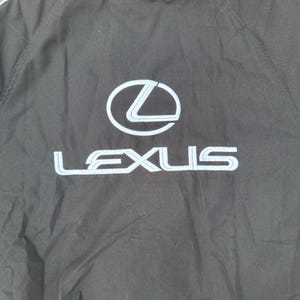 Pu&ograve; includere: Giacca nera con il logo Lexus bianco e azzurro sul retro. La giacca ha un cappuccio e una striscia azzurra lungo le spalle.