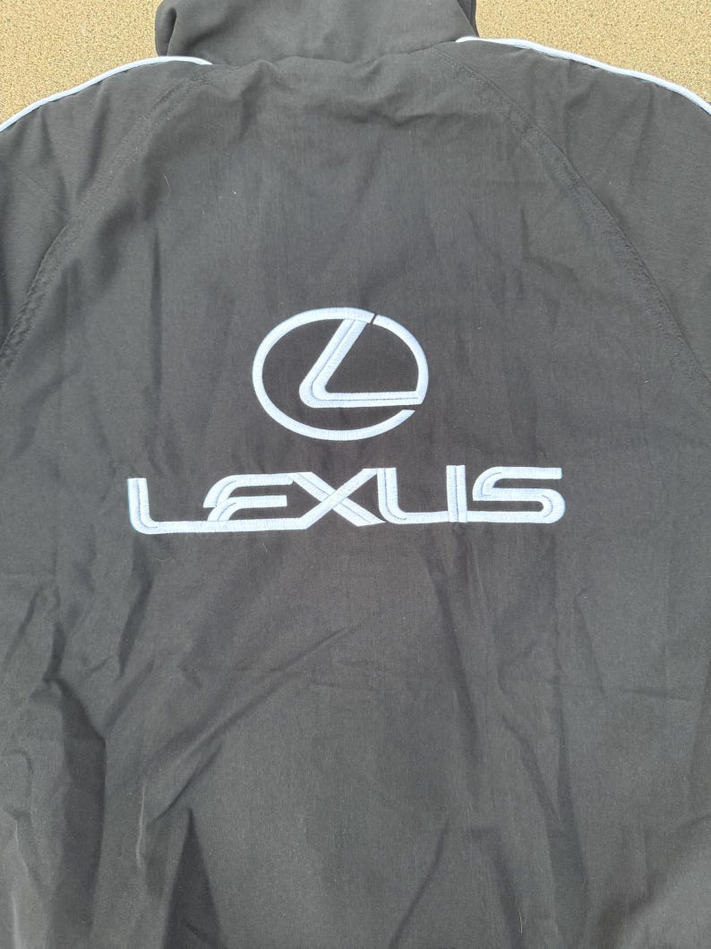Pu&ograve; includere: Giacca nera con il logo Lexus bianco e azzurro sul retro. La giacca ha un cappuccio e una striscia azzurra lungo le spalle.