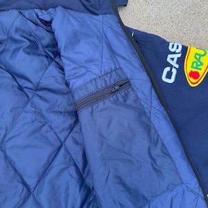 May include: Navy blue jacket with a quilted blue lining and a zippered pocket. The jacket features the text "CAS" in white and a yellow and red logo. The jacket has a dark blue exterior.