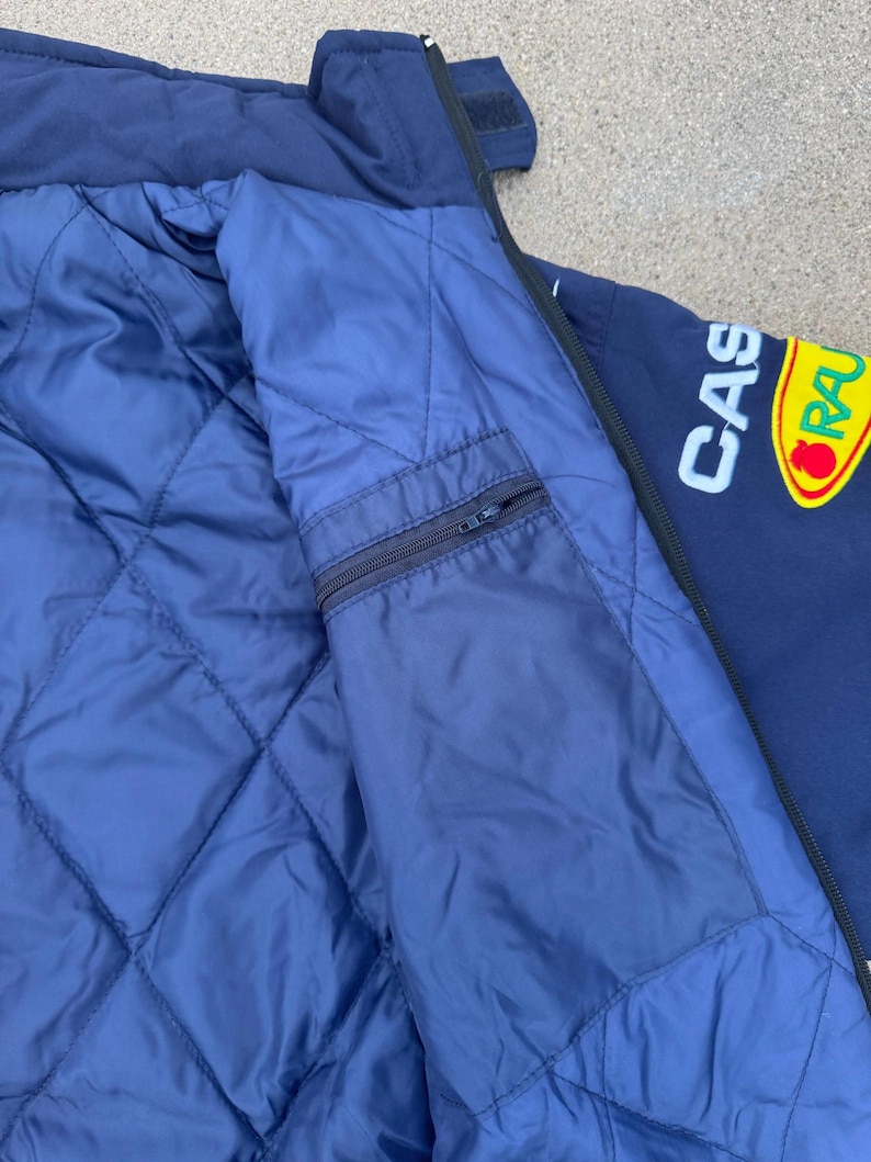 May include: Navy blue jacket with a quilted blue lining and a zippered pocket. The jacket features the text "CAS" in white and a yellow and red logo. The jacket has a dark blue exterior.
