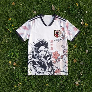 May include: A white V-neck short-sleeve jersey featuring a black and white graphic of an anime character, pink cherry blossom illustrations, and the Japan Football Association crest. The shirt includes black trim on the sleeves and collar.