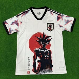 May include: A white V-neck soccer jersey featuring black trim on the collar and sleeves. The front displays a large red circle behind a black silhouette of a spiky-haired character, with the Japan Football Association crest and Japanese text.