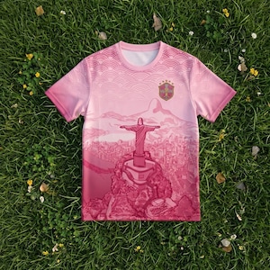 Könnte beinhalten: Ein kurzärmeliges T-Shirt in Rosatönen mit einem Grafikdruck der Christusstatue, die über eine Stadtlandschaft blickt. Der obere Teil zeigt ein Wellenmuster und auf der Brust befindet sich ein schildförmiges Emblem mit fünf Sternen.