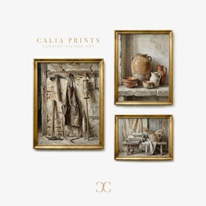 May include: Three framed vintage art prints with gold frames. The prints depict rustic scenes: a blacksmith's tools, pottery on a shelf, and a basket with folded textiles. The text "CALIA PRINTS CURATED VINTAGE ART" is visible.