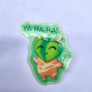May include: A sticker depicting a cartoon character with a green leaf-shaped face and a smiling expression. The character has an orange body with a spotted pattern and is holding a small stick. The text "YA HA HA!" is above the character.