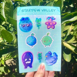 Peut inclure: Une planche d'autocollants Stardew Valley avec divers personnages et objets de dessins animés. Les autocollants incluent une myrtille avec des lunettes de soleil, une fleur violette, un fruit bleu, un fruit vert, un fantôme violet et un poulet bleu.