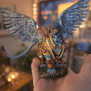 May include: A detailed figurine of a winged sphinx with a blue face, adorned with gold and red accents. The sculpture features intricate details on the wings and body, held in a hand. The figure has a blue tail and is set on a dark base.