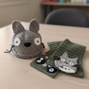 May include: A gray crocheted hat and scarf set featuring a cartoon cat design. The hat has large ears and white eyes, while the green scarf has cat and star appliqués. The set is handmade and suitable for children.