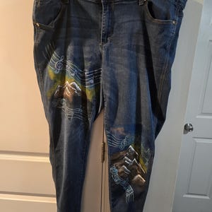 May include: Blue denim jeans with a hand-painted mountain and musical note design. The artwork features brown, green, and white accents. The jeans have a classic five-pocket design and a button and zipper closure.