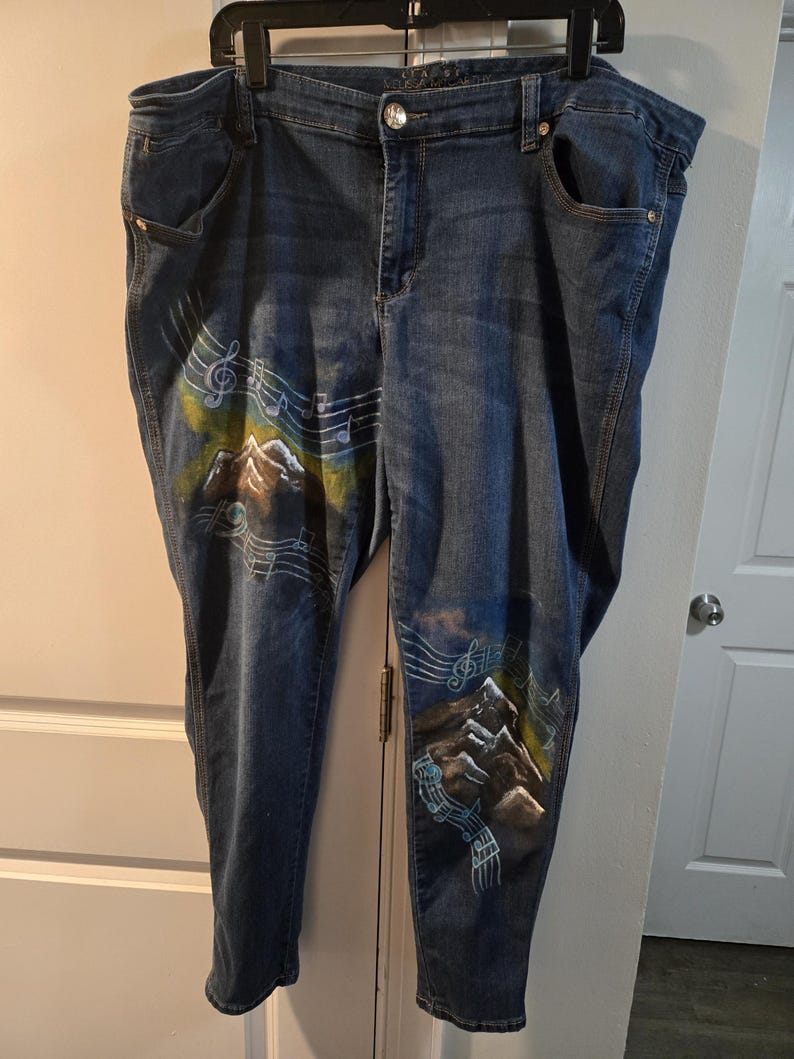 May include: Blue denim jeans with a hand-painted mountain and musical note design. The artwork features brown, green, and white accents. The jeans have a classic five-pocket design and a button and zipper closure.