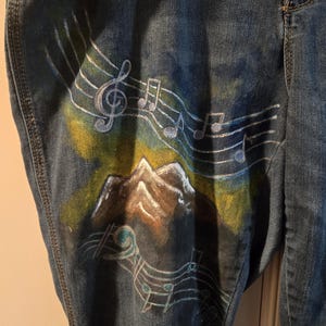 May include: Blue denim jeans with a hand-painted design featuring musical notes, a treble clef, and a mountain scene. The artwork incorporates shades of blue, white, yellow, and brown. The jeans have a classic five-pocket design.