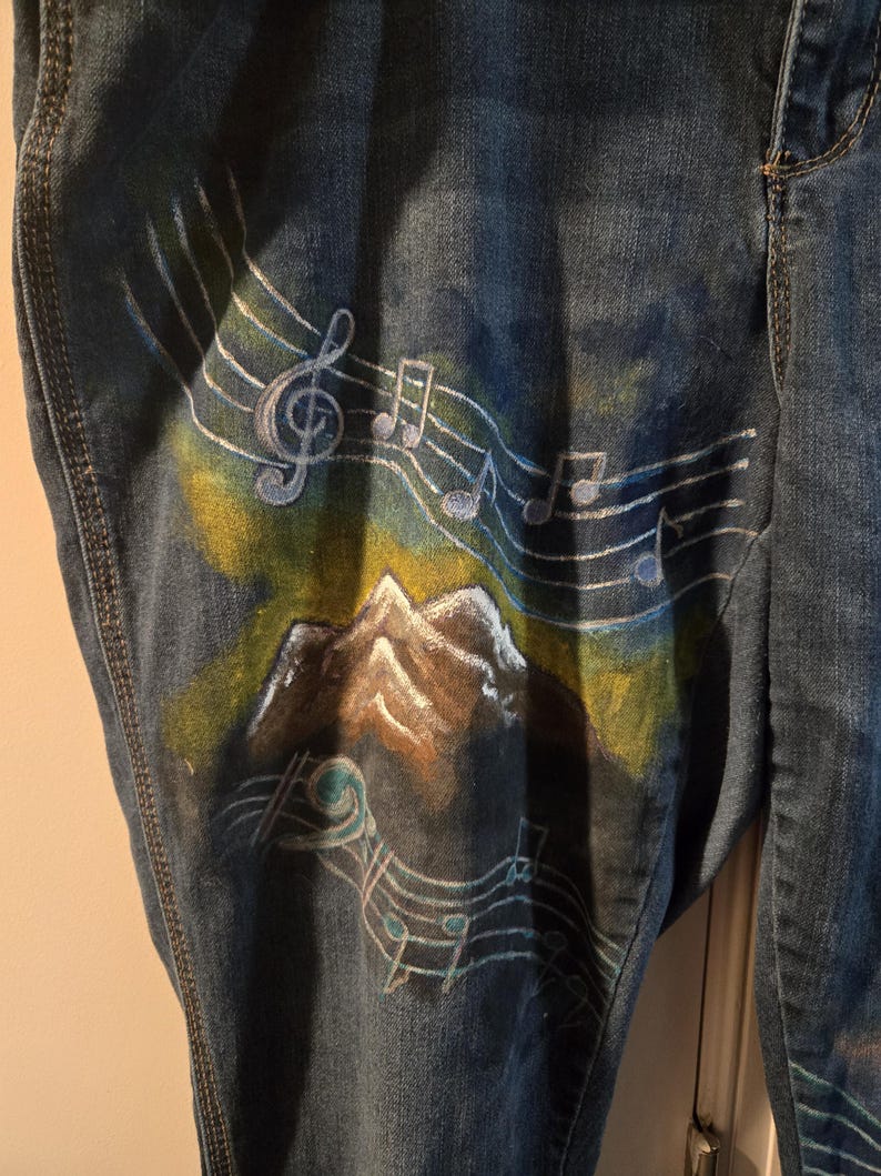 May include: Blue denim jeans with a hand-painted design featuring musical notes, a treble clef, and a mountain scene. The artwork incorporates shades of blue, white, yellow, and brown. The jeans have a classic five-pocket design.