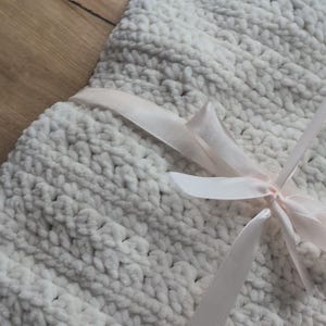 May include: A close-up of a cream-coloured, chunky knit blanket with a soft, textured appearance. The blanket is tied with a light pink satin ribbon. The edge of the blanket has a ruffled trim.