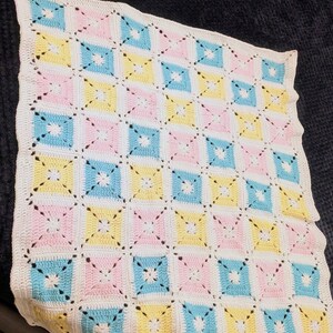 May include: A handmade crochet blanket with a repeating pattern of pastel squares in pink, yellow, and blue, separated by white crochet. The blanket has a scalloped white border and a delicate, openwork design.