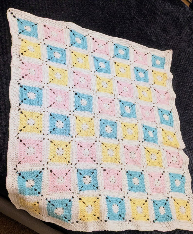 May include: A handmade crochet blanket with a repeating pattern of pastel squares in pink, yellow, and blue, separated by white crochet. The blanket has a scalloped white border and a delicate, openwork design.
