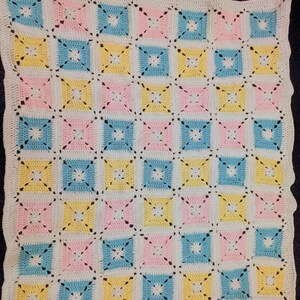 May include: A crocheted blanket with a repeating pattern of pastel squares in pink, yellow, and blue, separated by white and connected with black stitching. The blanket has a white scalloped border.