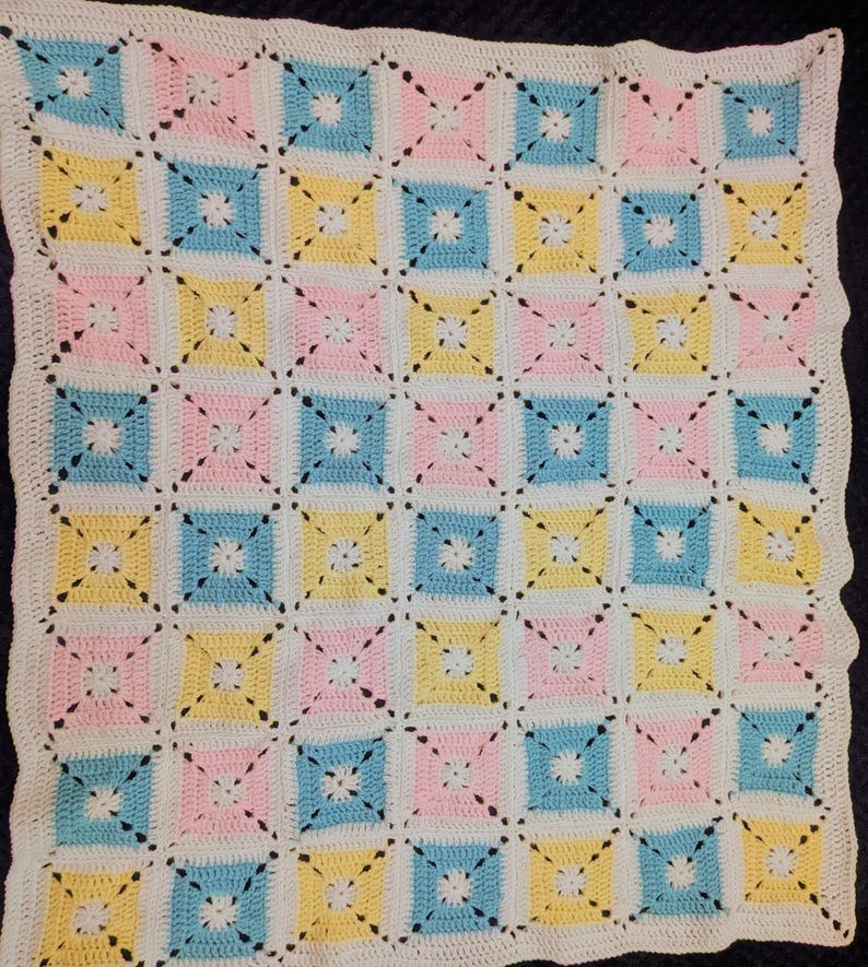 May include: A crocheted blanket with a repeating pattern of pastel squares in pink, yellow, and blue, separated by white and connected with black stitching. The blanket has a white scalloped border.