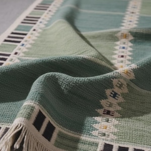 May include: A woven textile runner featuring a sage green and white color palette with a textured surface. The design includes geometric borders with black rectangular accents and a central white patterned stripe with small colorful details.