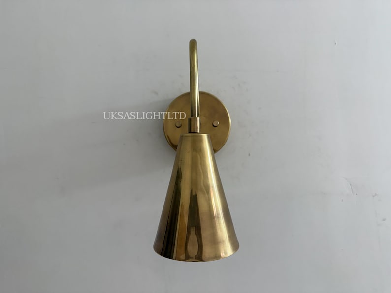 Puede incluir: Aplique de pared de lat&oacute;n con pantalla c&oacute;nica y brazo curvo. El aplique est&aacute; montado sobre una placa trasera circular. El texto "UKSASLIGHTLTD" es visible en la pared. Adecuado para iluminaci&oacute;n interior.