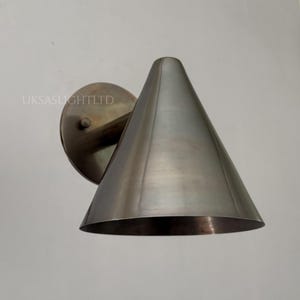 Pode incluir: Arandela de parede metálica com abajur cônico. A arandela tem uma base circular e um braço curvo, tudo em acabamento de metal escovado. A sombra é em forma de cone. Montado em uma parede branca.
