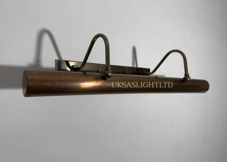Op de afbeelding: Een bronzen schilderijlamp met een cilindrisch lichaam en gebogen armen. Op de lamp staat de tekst "UKSASLIGHTLTD" gedrukt. De lamp is gemonteerd op een rechthoekige basis.
