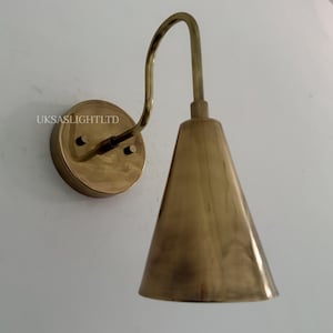 Puede incluir: Aplique de pared de lat&oacute;n con un brazo curvo y una pantalla c&oacute;nica. El aplique est&aacute; montado sobre una base redonda y tiene un acabado met&aacute;lico c&aacute;lido. El texto "UKSASLIGHTLTD" es visible en la base.