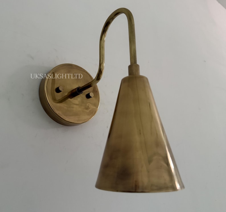 Puede incluir: Aplique de pared de lat&oacute;n con un brazo curvo y una pantalla c&oacute;nica. El aplique est&aacute; montado sobre una base redonda y tiene un acabado met&aacute;lico c&aacute;lido. El texto "UKSASLIGHTLTD" es visible en la base.