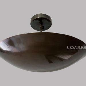 Könnte beinhalten: Eine moderne Deckenleuchte in Bronze. Die Leuchte hat einen großen, flachen, ovalen Schirm und eine kreisförmige Montageplatte. Sie wird von einer kurzen Stange von der Decke abgehängt. Die Oberfläche des Schirms ist reflektierend.