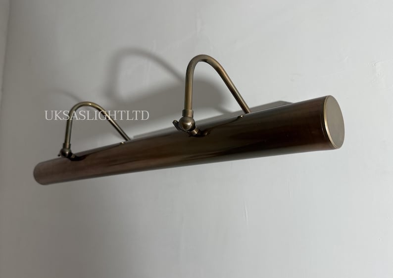 Op de afbeelding: Een bronzen schilderijverlichting met twee gebogen armen en een cilindrisch lichaam. De armatuur is aan een witte muur bevestigd. De tekst "UKSASLIGHTLTD" is zichtbaar.