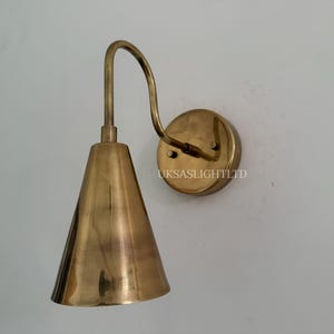 Puede incluir: Aplique de pared de lat&oacute;n con pantalla c&oacute;nica y brazo curvo. El aplique tiene una base circular y est&aacute; montado en una pared blanca. El texto "UKSASLIGHTLTD" es visible en la base.