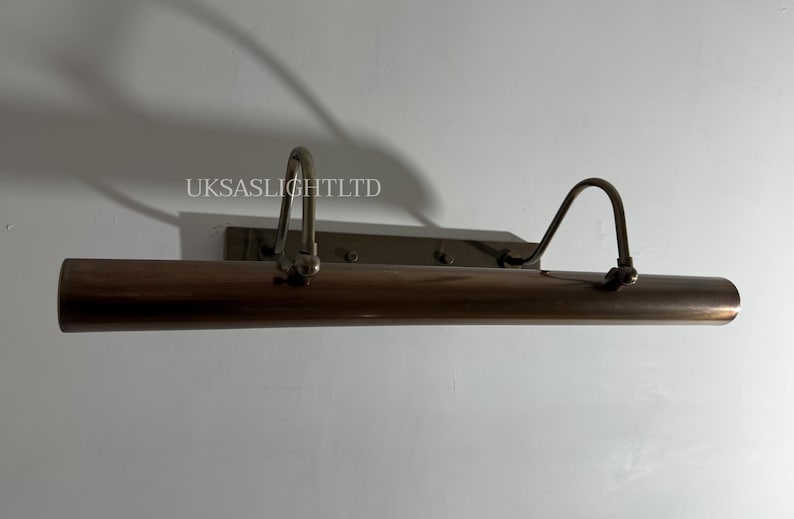 Op de afbeelding: Een bronzen schilderijlamp met twee gebogen armen en een cilindrische lichtbehuizing. De armatuur is gemonteerd op een rechthoekige achterplaat. De tekst "UKSASLIGHTLTD" is zichtbaar.