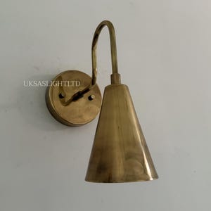 Puede incluir: Una l&aacute;mpara de pared de lat&oacute;n con una pantalla c&oacute;nica y un brazo curvo. La l&aacute;mpara tiene una base circular y est&aacute; montada en una pared blanca. El texto "UKSASLIGHTLTD" es visible en la base.