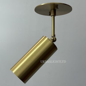 May include: A brass-colored, cylindrical spotlight fixture with a circular ceiling mount. The spotlight is angled downward and features the text "UKSASLIGHTLTD" on its side. The fixture is designed for directional lighting.