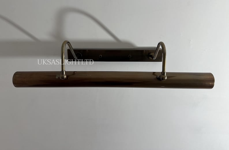 Op de afbeelding: Een bronzen schilderijverlichting met twee gebogen armen en een rechthoekige achterplaat. De lamp is aan een witte muur bevestigd. De tekst "UKSASLIGHTLTD" is zichtbaar.