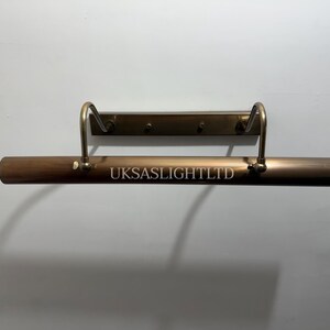 Op de afbeelding: Een bronzen fotolamp met een rechthoekige balk en gebogen armen. De tekst "UKSASLIGHTLTD" is op de balk gedrukt. De lamp is aan een muur bevestigd.