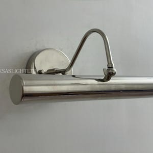May include: A polished nickel picture light fixture. The light has a cylindrical body with a curved arm and a round wall mount. The fixture is designed to illuminate artwork or other wall-mounted items. The text "UKSASLIGHTLTD" is visible.