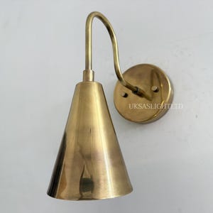 Puede incluir: Aplique de pared de lat&oacute;n con una pantalla c&oacute;nica y un brazo curvo. El aplique tiene un soporte de pared circular y un acabado met&aacute;lico c&aacute;lido. El texto "UKSASLIGHTLTD" es visible en el soporte de pared.