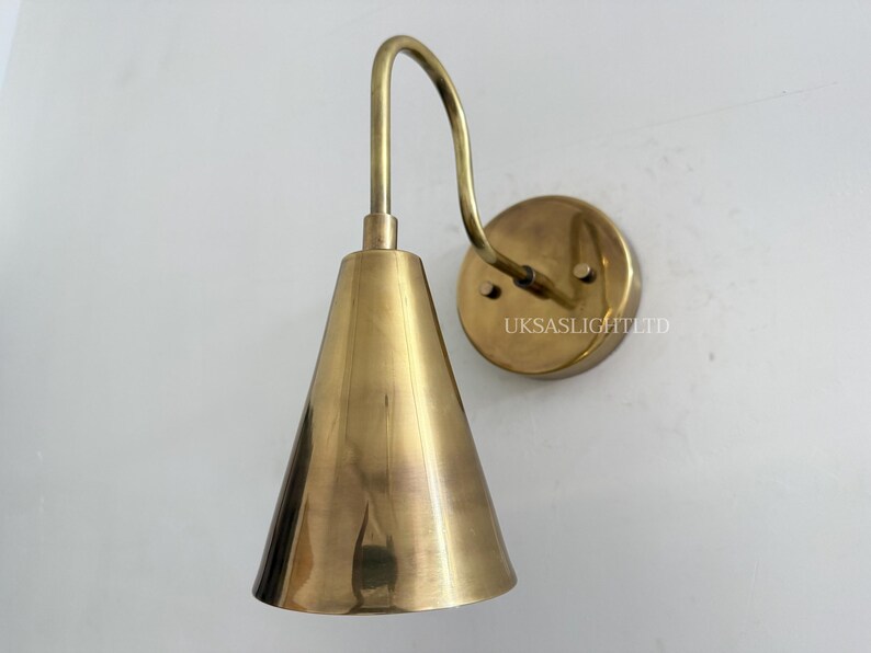 Puede incluir: Aplique de pared de lat&oacute;n con una pantalla c&oacute;nica y un brazo curvo. El aplique tiene un soporte de pared circular y un acabado met&aacute;lico c&aacute;lido. El texto "UKSASLIGHTLTD" es visible en el soporte de pared.