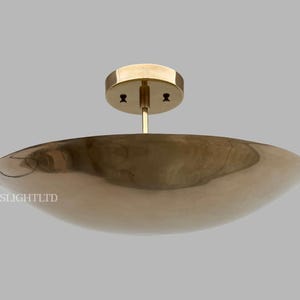 May include: A modern, brass-colored ceiling light fixture. The fixture has a shallow, bowl-shaped shade and a circular mounting plate. The design is minimalist and elegant, suitable for various interior styles. The fixture is suspended from a central rod.