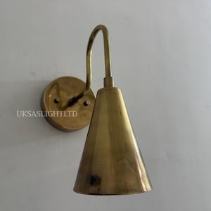 Puede incluir: Aplique de pared de lat&oacute;n con pantalla c&oacute;nica y brazo curvo. El aplique tiene una base redonda y est&aacute; montado en una pared blanca. El texto "UKSASLIGHTLTD" es visible en la base. Adecuado para iluminaci&oacute;n interior.