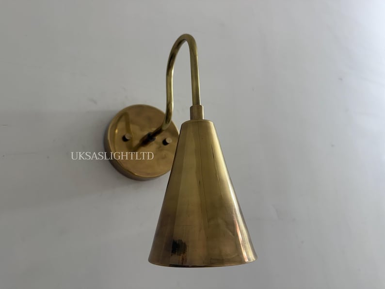 Puede incluir: Aplique de pared de lat&oacute;n con pantalla c&oacute;nica y brazo curvo. El aplique tiene una base redonda y est&aacute; montado en una pared blanca. El texto "UKSASLIGHTLTD" es visible en la base. Adecuado para iluminaci&oacute;n interior.