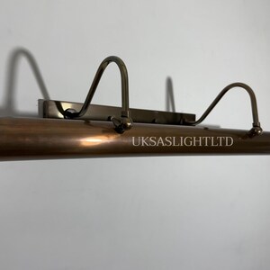Op de afbeelding: Een bronzen schilderijlamp met een cilindrisch lichaam en gebogen armen. Op de lamp staat de tekst "UKSASLIGHTLTD" gedrukt. De lamp is gemonteerd op een rechthoekige basis.