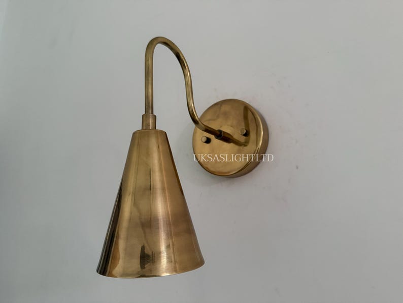 Puede incluir: Aplique de pared de lat&oacute;n con pantalla c&oacute;nica y brazo curvo. El aplique tiene una base circular y est&aacute; montado en una pared blanca. El texto "UKSASLIGHTLTD" est&aacute; impreso en la base.