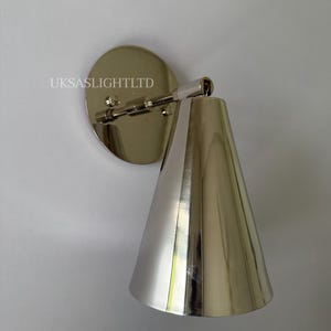 Op de afbeelding: Zilveren wandlamp met een conische kap en een cirkelvormige achterplaat. De lamp is aan een witte muur bevestigd. De tekst "UKSASLIGHTLTD" staat op de achterplaat.