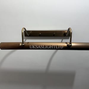 Op de afbeelding: Een bronzen fotolamp met een rechthoekige balk en gebogen armen. De tekst "UKSASLIGHTLTD" is op de balk gedrukt. De lamp is aan een muur bevestigd.
